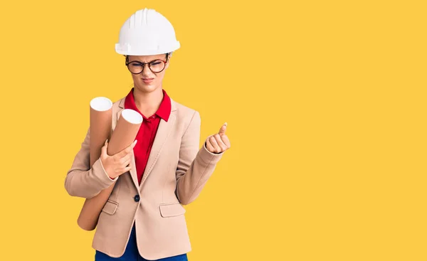 Young beautiful woman wearing architect hardhat and glasses holding blueprints annoyed and frustrated shouting with anger, yelling crazy with anger and hand raised