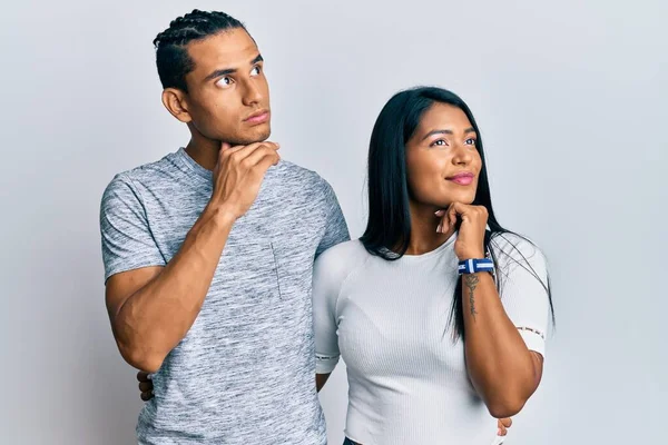 若いラテン系のカップルの質問 集中的な表現について考える顎に手でカジュアルな服を着て 思慮深い顔で笑顔 疑わしい概念 — ストック写真