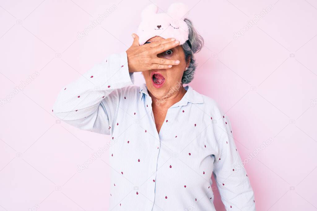Senior hispanic grey- haired woman wearing sleep mask and pajama peeking in shock covering face and eyes with hand, looking through fingers afraid 