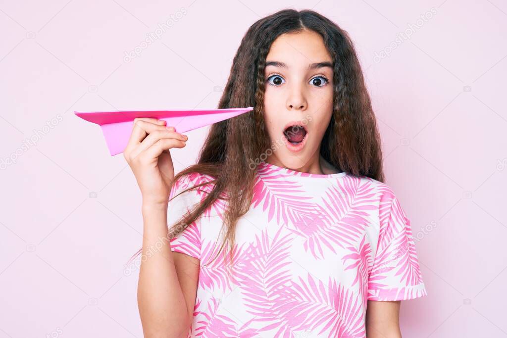 Cute hispanic child girl holding paper airplane scared and amazed with open mouth for surprise, disbelief face 