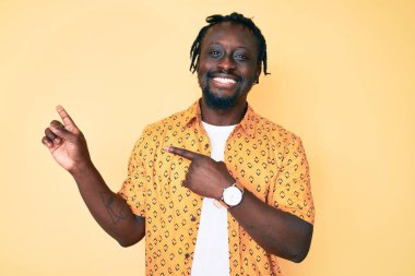 Saçları örgülü, gülümseyen ve kameraya bakıp iki eliyle ve parmaklarıyla yüzünü gösteren Afro-Amerikan genç adam.. 