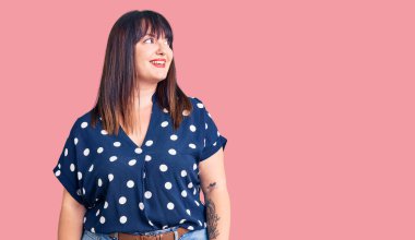 Genç ve cüsseli bir kadın, sıradan kıyafetler giyiyor. Yüzünde gülümsemeyle yan yana bakıyor, doğal bir ifade. Kendinden emin gülen. 