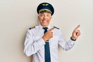 Yakışıklı, orta yaşlı, olgun, uçak pilotu üniforması giyen, gülümseyen ve kameraya bakan iki eliyle ve parmaklarıyla yan tarafı gösteren bir adam.. 