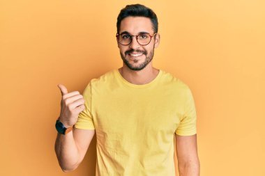Gündelik kıyafetler ve gözlük takan, gülen yüzlü gülümseyen ve başparmağıyla yan tarafı gösteren İspanyol genç adam.. 