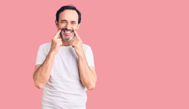 Orta yaşlı yakışıklı bir adam günlük tişört giyiyor ağzı açık gülümsüyor, parmakları işaret ediyor ve zorla neşeli bir gülümseme sergiliyor. 