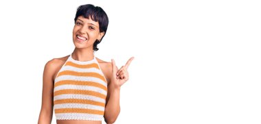 Kısa saçlı, sıradan kıyafetler giyen, yüzünde kocaman bir gülümseme olan, eli kameraya bakan esmer bir kadın.. 