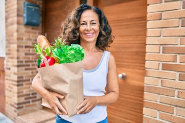 Orta yaşlı İspanyol kadın, elinde alışveriş torbalarıyla şehirde mutlu bir şekilde gülümsüyor..