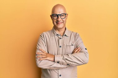 Orta yaşlı, kel, sıradan kıyafetler ve gözlüklü mutlu yüzlü, kollarını kavuşturup kameraya bakan bir adam. pozitif kişi. 