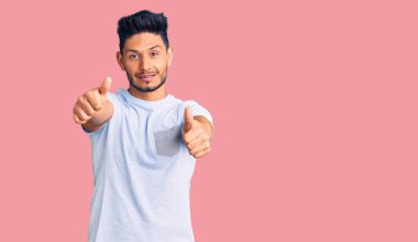 Yakışıklı Latin Amerikalı genç adam gündelik giysiler giyip, el hareketleriyle olumlu jest yapıyor, gülümsüyor ve başarı için seviniyor. kazanan jest. 