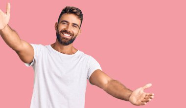 Latin asıllı genç bir adam, günlük beyaz tişört giyip kameraya bakıyor ve sarılmak için kollarını açmış gülümsüyor. Mutluluğu kucaklayan neşeli bir ifade.. 