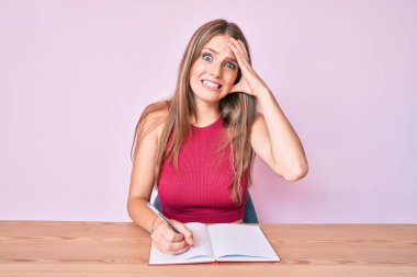 Masaya oturmuş not defteri yazan beyaz bir kız. Elleri başının üstünde, şaşırmış ve sinirli bir halde. 