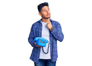 Yakışıklı Latin Amerikalı genç adam elinde eski bir telefon, ciddi bir yüz, eli çenesinde soruyu düşünüyor, kafa karıştırıcı bir fikir. 