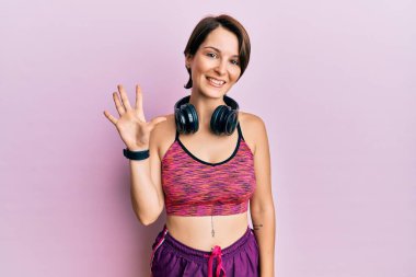 Kısa saçlı, spor giyimli, kulaklık takan genç esmer kadın beş numarayı gösterip işaret ederken kendinden emin ve mutlu bir şekilde gülümsüyor.. 
