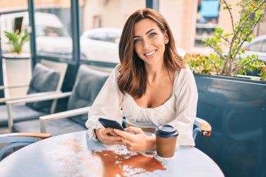 Genç İspanyol kadın kahvehanenin terasında akıllı telefon kullanarak mutlu gülümsüyor..