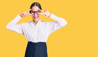 Güzel, esmer, profesyonel garson önlüğü giymiş, yüzünde parmaklarıyla barış sembolü çizen, neşeli, zaferi gösteren genç bir kadın. 