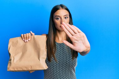 Young hispanic woman holding take away paper bag with open hand doing stop sign with serious and confident expression, defense gesture 