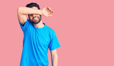 Sakallı, sıradan tişörtler giyen, gözleri kollarıyla kaplayan neşeli ve komik gülümseyen genç yakışıklı bir adam. kör kavram. 