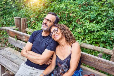 Orta yaşlı çift gökyüzüne bakarken mutlu bir şekilde gülümsüyor. Parkta chihuahua köpeğiyle bankta oturmak.