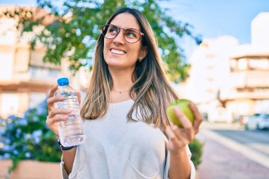 Genç beyaz kadın gülümsüyor. Şehirde yeşil elma ve bir şişe su tutuyor..