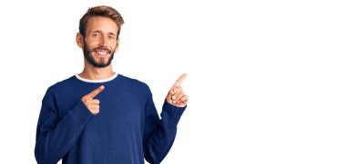 Sakallı, sıradan bir süveter giyen yakışıklı sarışın. Gülümseyen ve kameraya bakan. İki eli ve parmakları yanda olan.. 