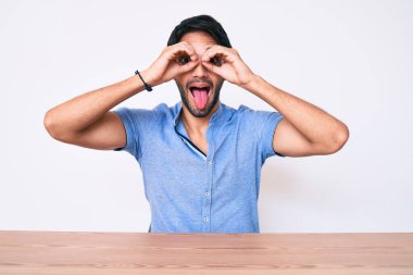 Masada oturan, sıradan kıyafetler giyen yakışıklı İspanyol bir adam. Dürbün gibi jest yapıyor, gözleri parmaklarından bakıyor. çılgın ifade. 