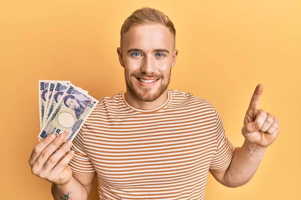 Young caucasian man holding japanese yen banknotes smiling with an idea or question pointing finger with happy face, number one 