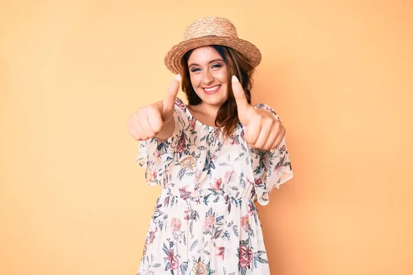 若い美しい白人女性は夏のドレスを着て 帽子は手で肯定的なジェスチャーを行うことを承認し 親指を上げて笑顔と成功のために幸せ 勝者のジェスチャー — ストック写真