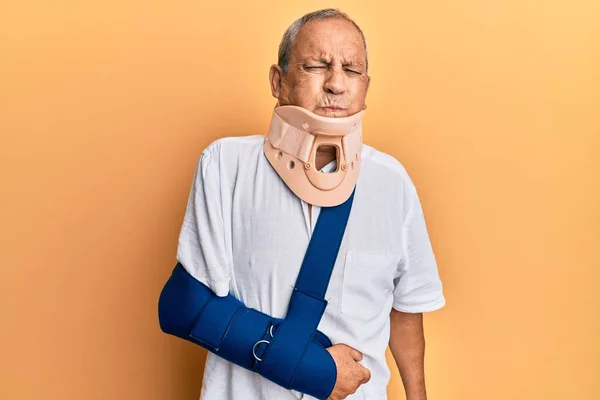 Handsome mature senior man wearing cervical collar and arm on sling puffing cheeks with funny face. mouth inflated with air, crazy expression.