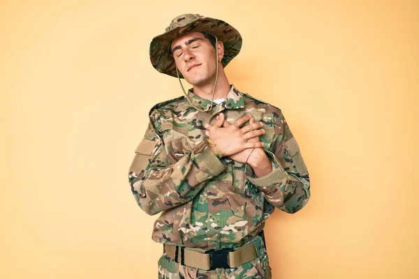 目を閉じて胸の上で手で笑みを浮かべてカモフラージュ軍の制服を着て若いハンサムな男と顔に感謝のジェスチャー 健康の概念 — ストック写真