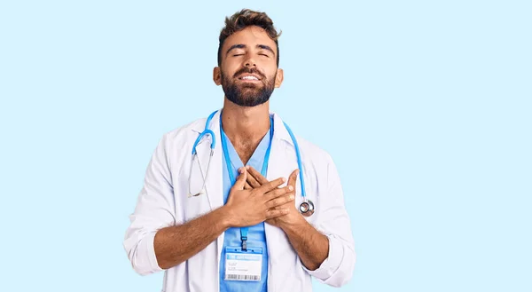 身穿医生制服的年轻人 戴着听诊器 双手放在胸前笑着 闭着眼睛 脸上挂着感激的手势 健康概念 — 图库照片