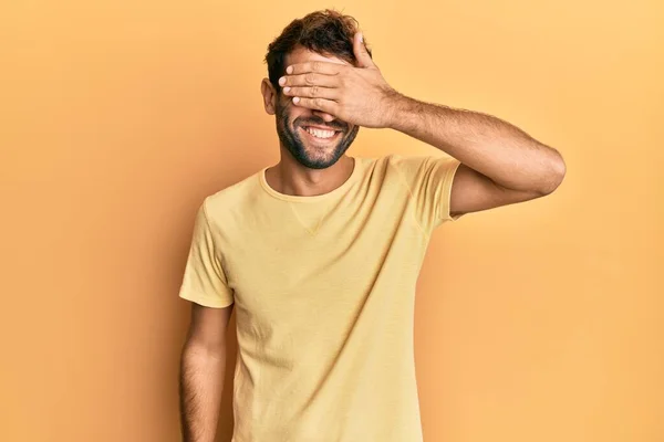 黄色の背景にカジュアルな黄色のTシャツを着て髭を生やしたハンサムな男は笑顔で 驚きのために目を覆う顔を手で笑っています 視覚障害の概念 — ストック写真