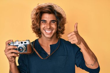 İspanyol asıllı genç adam elinde eski model bir kamera, gülümsüyor mutlu ve pozitif, başparmağıyla mükemmel ve onay işareti yapıyor. 