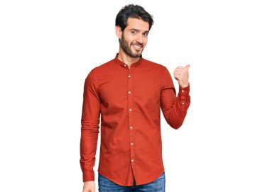 Gündelik kıyafetler giyen, gülümseyen, gülümseyen ve başparmağıyla yan tarafı gösteren İspanyol genç adam.. 