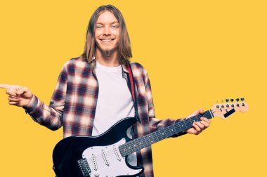 Uzun saçlı yakışıklı beyaz adam elektro gitar çalıyor. Gülümsüyor, mutlu bir şekilde elini ve parmağını yana doğru gösteriyor. 