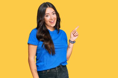 Güzel, esmer, sıradan kıyafetler giyen, yüzünde kocaman bir gülümseme olan genç bir kadın, kameraya el ile işaret ediyor.. 