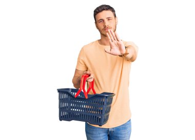 Elinde ayı süpermarketi olan yakışıklı genç adam elinde alışveriş sepetiyle dur işareti yapıyor ciddi ve kendinden emin bir ifadeyle, savunma jesti yapıyor. 