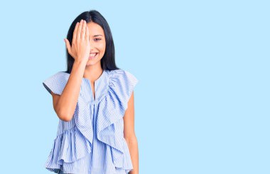 Genç, güzel, latin elbiseli bir kız bir gözünü eliyle kapatıyor, yüzünde kendine güvenen bir gülümseme ve sürpriz bir duygu var.. 