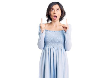 Gündelik giysiler giyen genç ve güzel bir kız şaşırmış ve şaşırmış bir şekilde yukarı bakıyor ve parmaklarıyla işaret ediyor ve kollarını kaldırıyor.. 