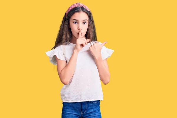 サイドに手を指差して唇に指で静かになるように頼むカジュアルな白いTシャツを着てかわいいヒスパニック系の子供の女の子 沈黙と秘密の概念 — ストック写真