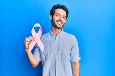 Genç İspanyol adam pembe kanser kurdelesi tutuyor. Pozitif ve mutlu görünüyor. Kendine güvenen bir gülümsemeyle dişlerini gösteriyor. 