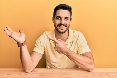 Gündelik kıyafetler giyen genç İspanyol adam masaya oturmuş kameraya hayret ediyor ve gülümsüyor.. 