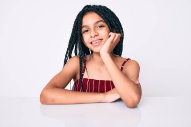 Saçları örgülü, neşeli ve havalı bir gülümsemeyle masada oturan Afro-Amerikan kız çocuğu. şanslı kişi. 