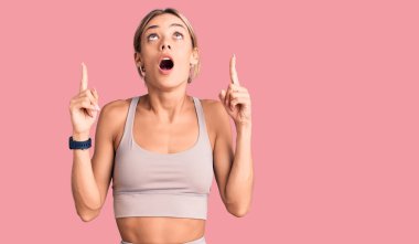 Spor kıyafetleri giyen güzel beyaz kadın şaşırmış ve şaşırmış bir şekilde yukarı bakıyor ve parmaklarıyla işaret ediyor ve kolunu kaldırıyor.. 