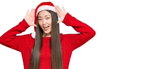 若い中国の女性は陽気に手を顔を示すと竹をのぞき見再生笑顔クリスマス帽子を身に着けている びっくりして興奮しました — ストック写真