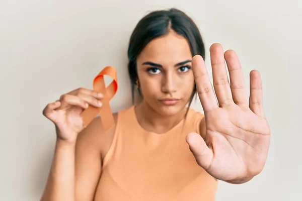 Young brunette woman holding awareness orange ribbon with open hand doing stop sign with serious and confident expression, defense gesture