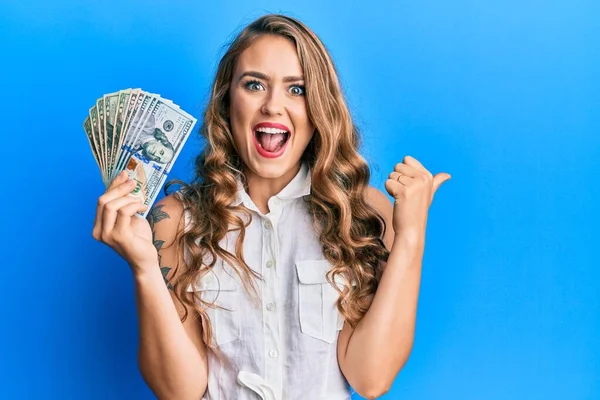 Joven Chica Rubia Sosteniendo Dólares Apuntando Pulgar Hacia Lado Sonriendo — Foto de Stock