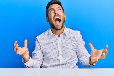 İş kıyafetleri giymiş yakışıklı bir İspanyol masaya oturmuş deli gibi bağırıyor, bağırıyor ve agresif bir ifadeyle kollarını kaldırıyor. hüsran kavramı. 