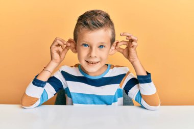 Kafkasyalı sevimli bir çocuk, günlük kıyafetler giymiş masaya oturmuş gülümsüyor kulakları parmaklarıyla çekiyor, komik bir jest. deneme sorunu 