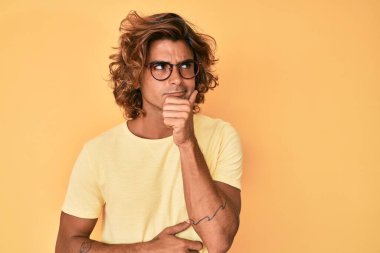 Gündelik kıyafetler ve gözlük takan genç İspanyol bir adam ciddi bir yüz ifadesi, eli çenesinde soru soran, kafa karıştırıcı bir fikir hakkında düşünceli. 