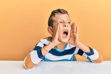Beyaz elbiseli sevimli bir çocuk masada oturmuş elleri ağızda bağırarak bağırıyor. 